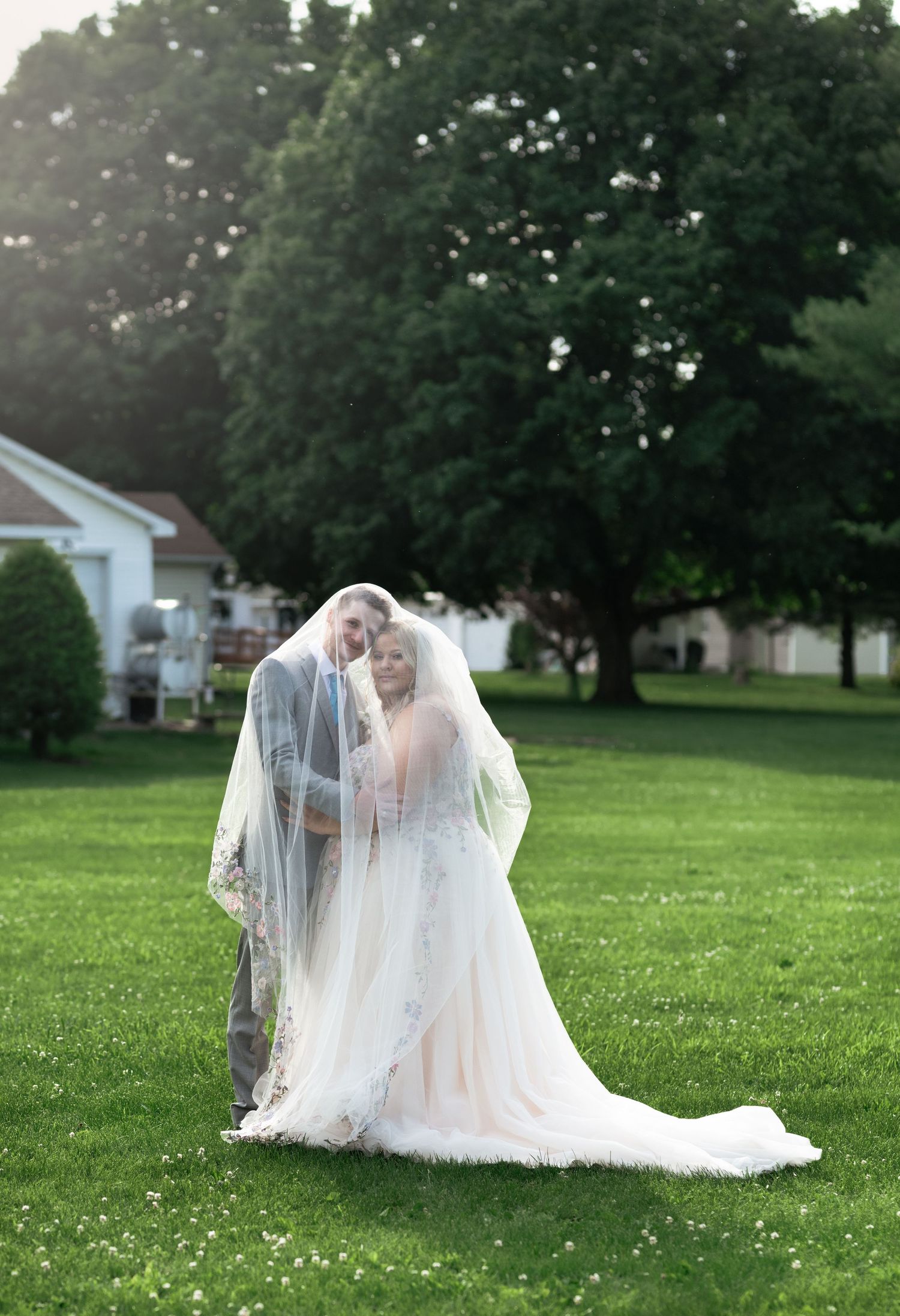 Bride and groom share a romantic moment under a flowing veil on a lush green lawn with trees in the background.
