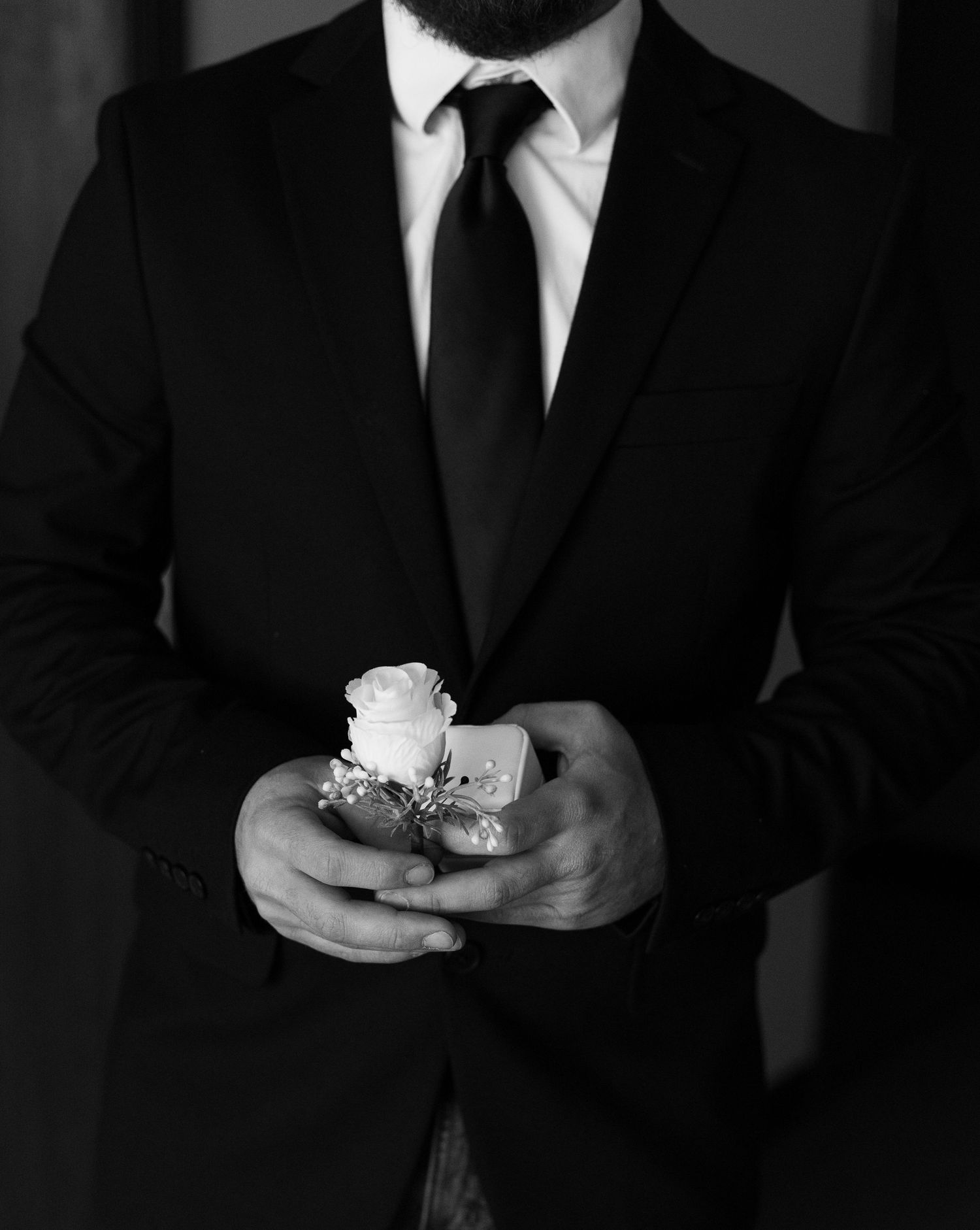 Black and white photo of a groom in a suit holding a white rose boutonniere before his wedding ceremony.