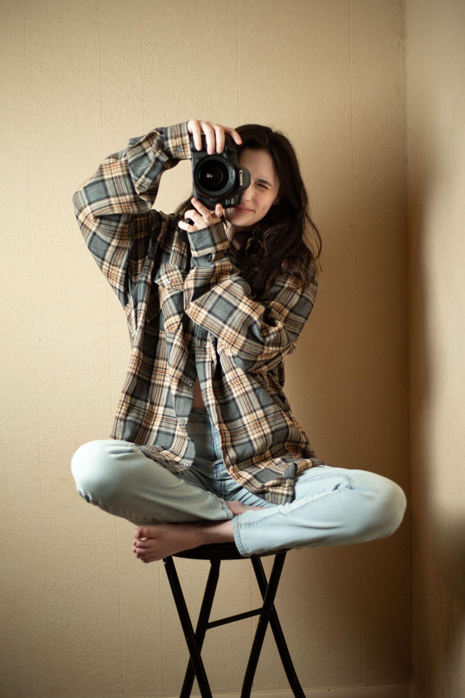 Young woman in plaid shirt sits cross-legged on stool, holding DSLR camera up to her face, photographing viewer.