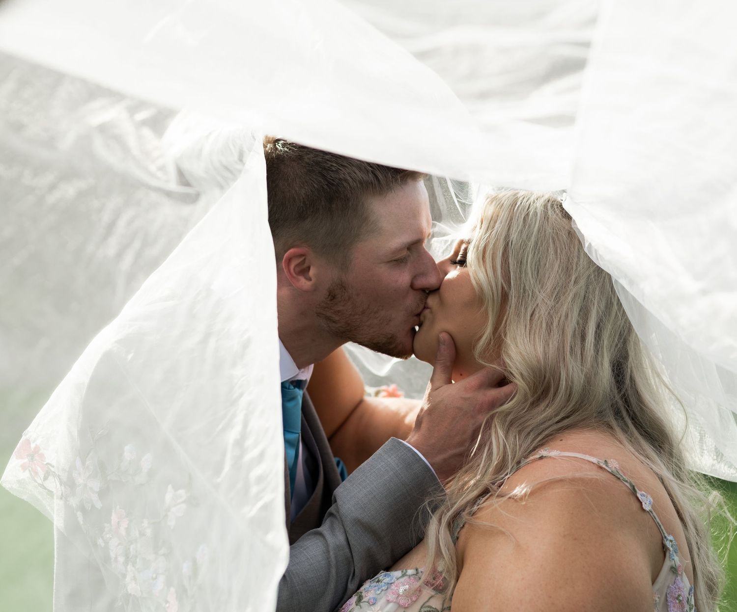 Bride and groom sharing a romantic kiss under a flowing white wedding veil outdoors.