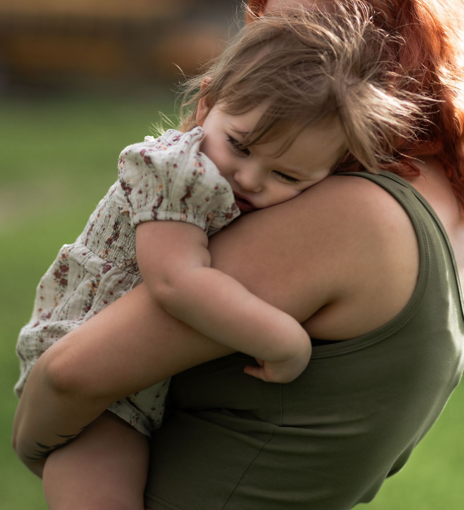 A mother with red hair tenderly holds a sleeping toddler girl in a floral dress against her shoulder outdoors.
