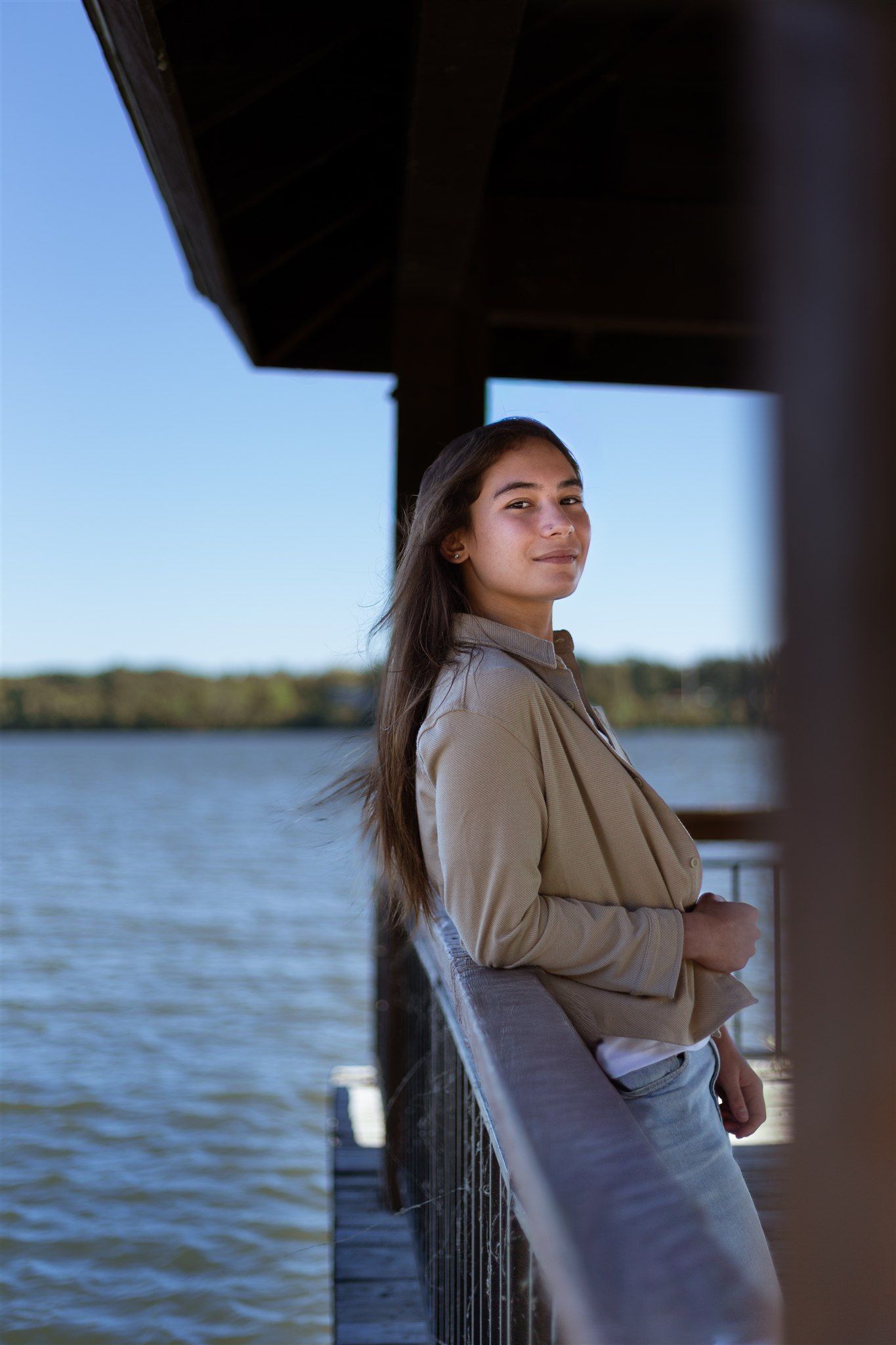Woman in beige jacket poses against railing overlooking serene lake at sunset.