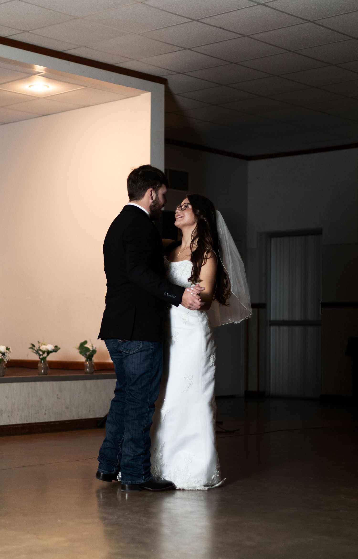 A couple shares their first dance at their wedding reception in a dimly lit room with white walls.