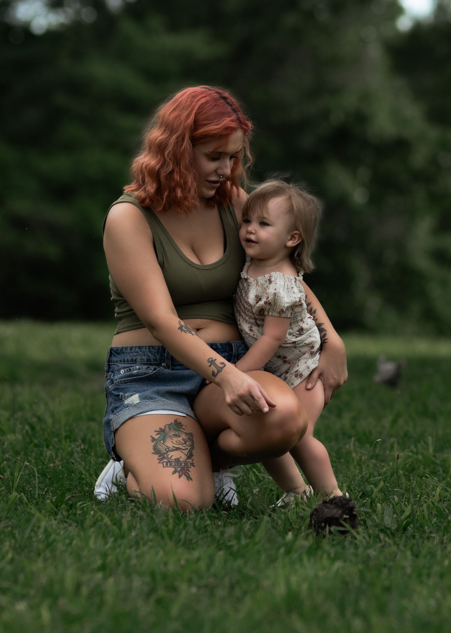 Red-haired tattooed mother crouching in green grass park, holding toddler child in floral outfit on sunny day.
