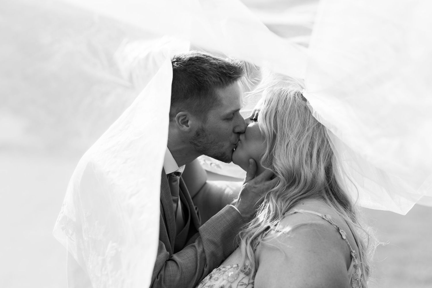 Black and white wedding photo of bride and groom kissing under flowing veil.