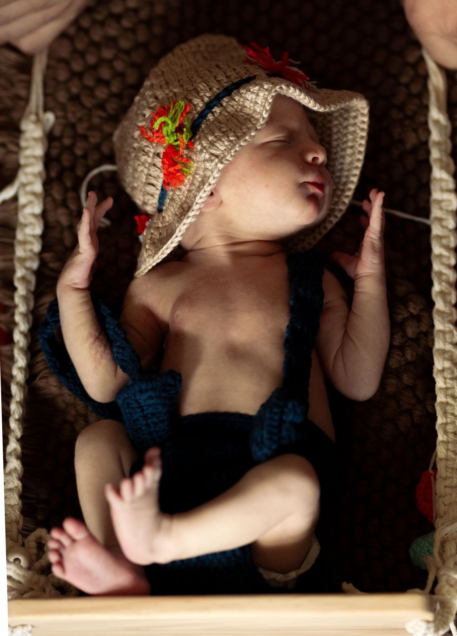 Newborn baby sleeping in a crochet hammock wearing a knit hat with floral accents and blue suspenders.