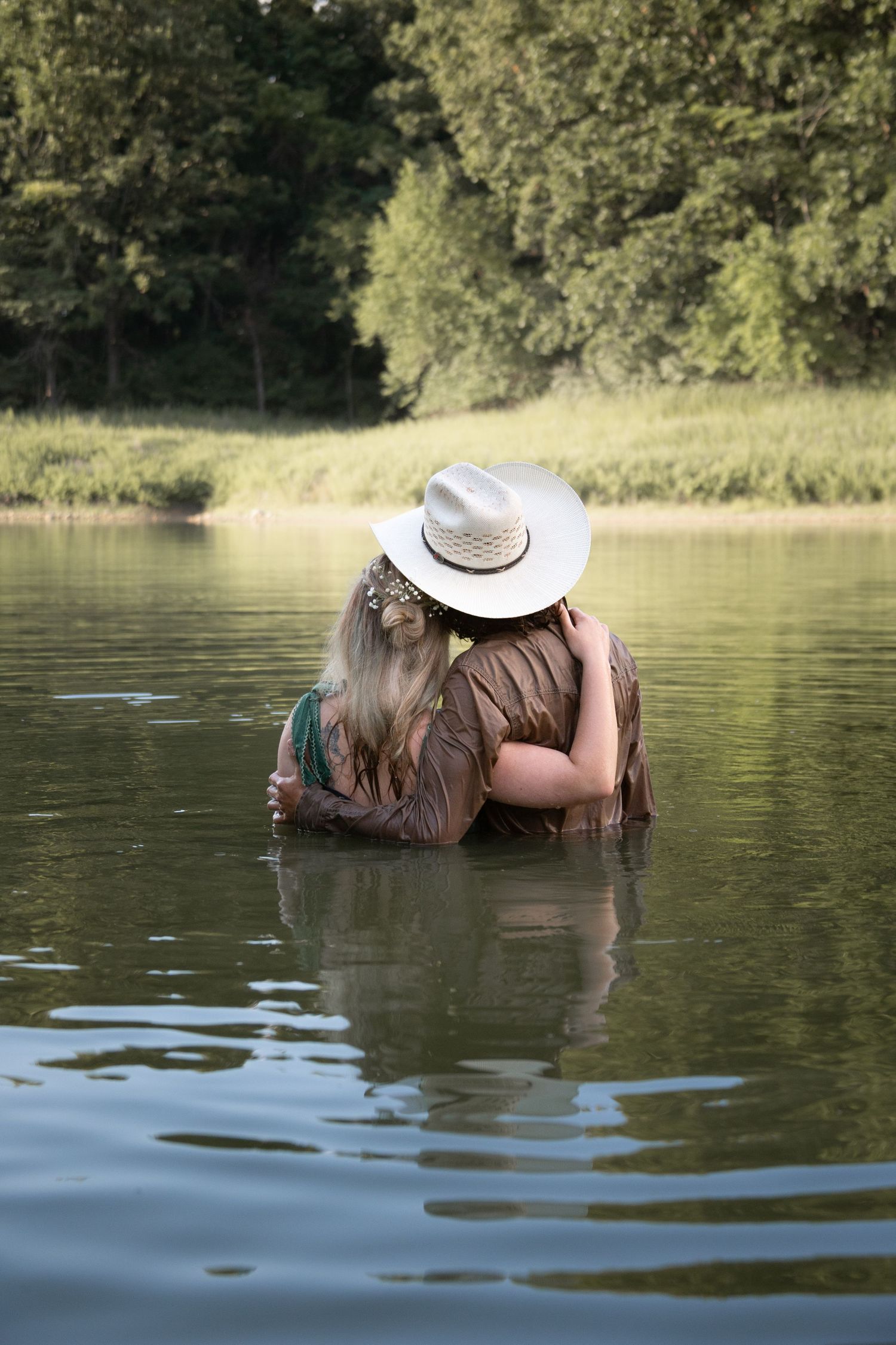 A couple embraces while wading in a serene lake surrounded by trees on a sunny day.