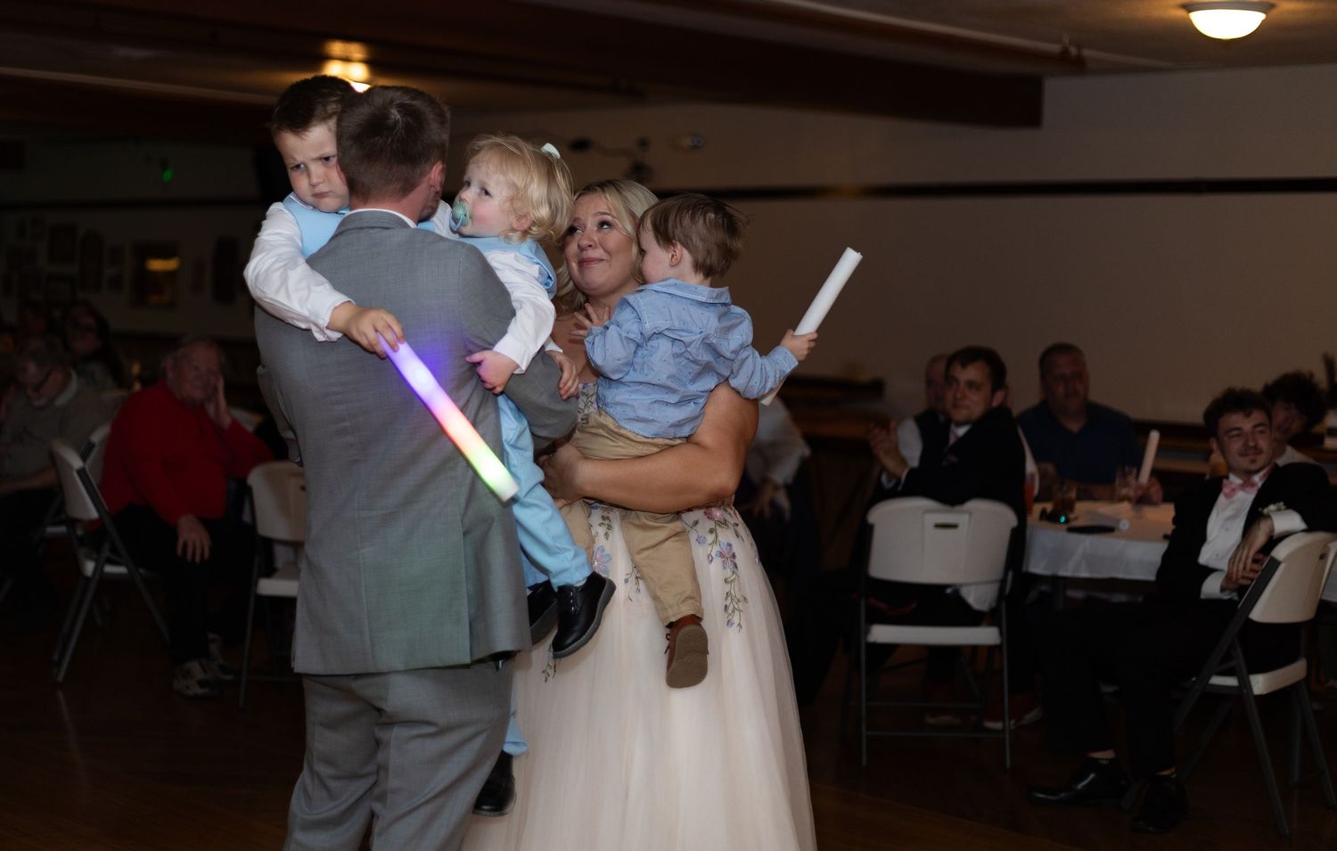 Bride and groom dancing with two young children at a wedding reception, holding glow sticks on the dance floor.