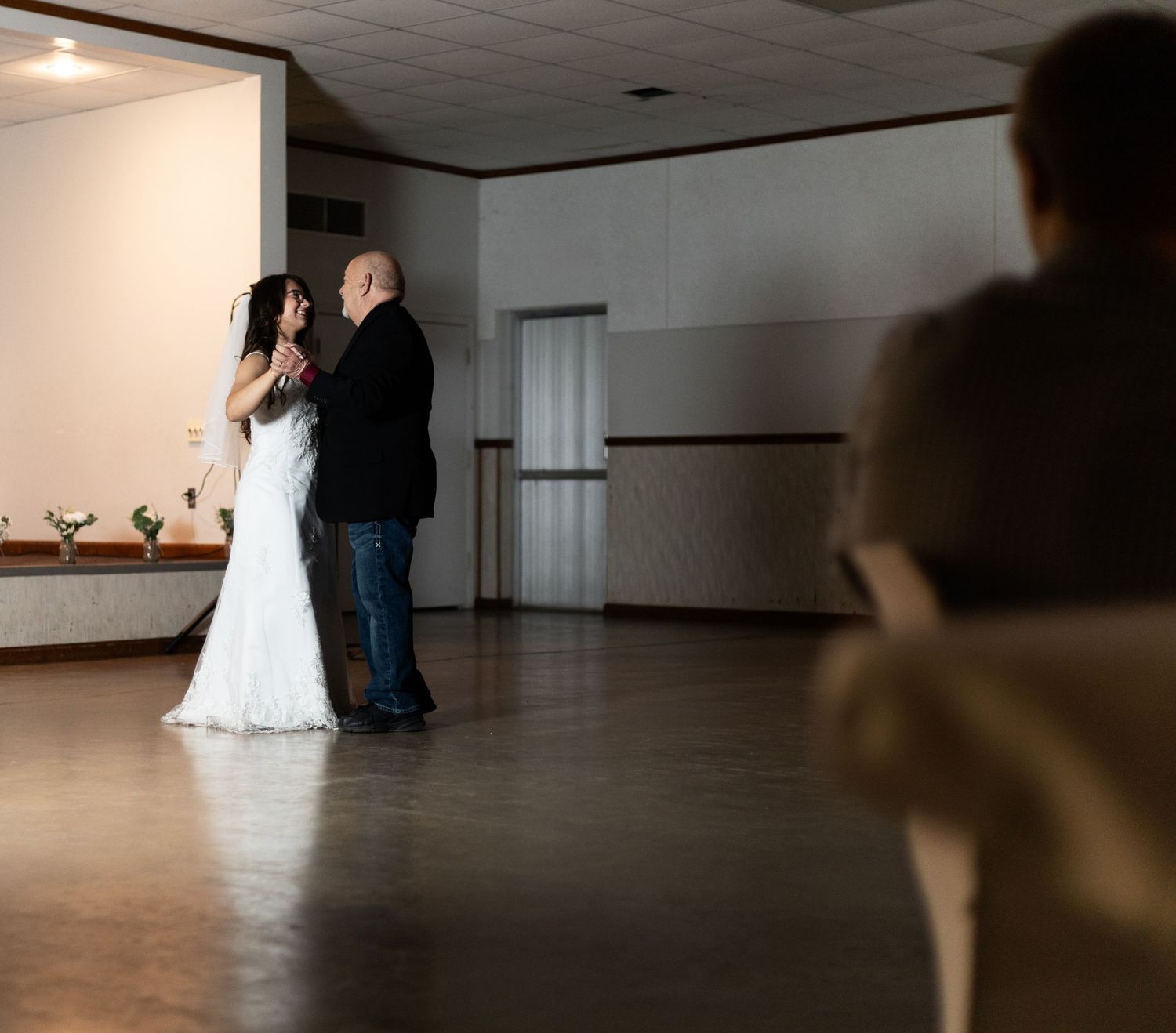 A couple in formal wedding attire dance together in an empty studio with a mirror-lined wall.