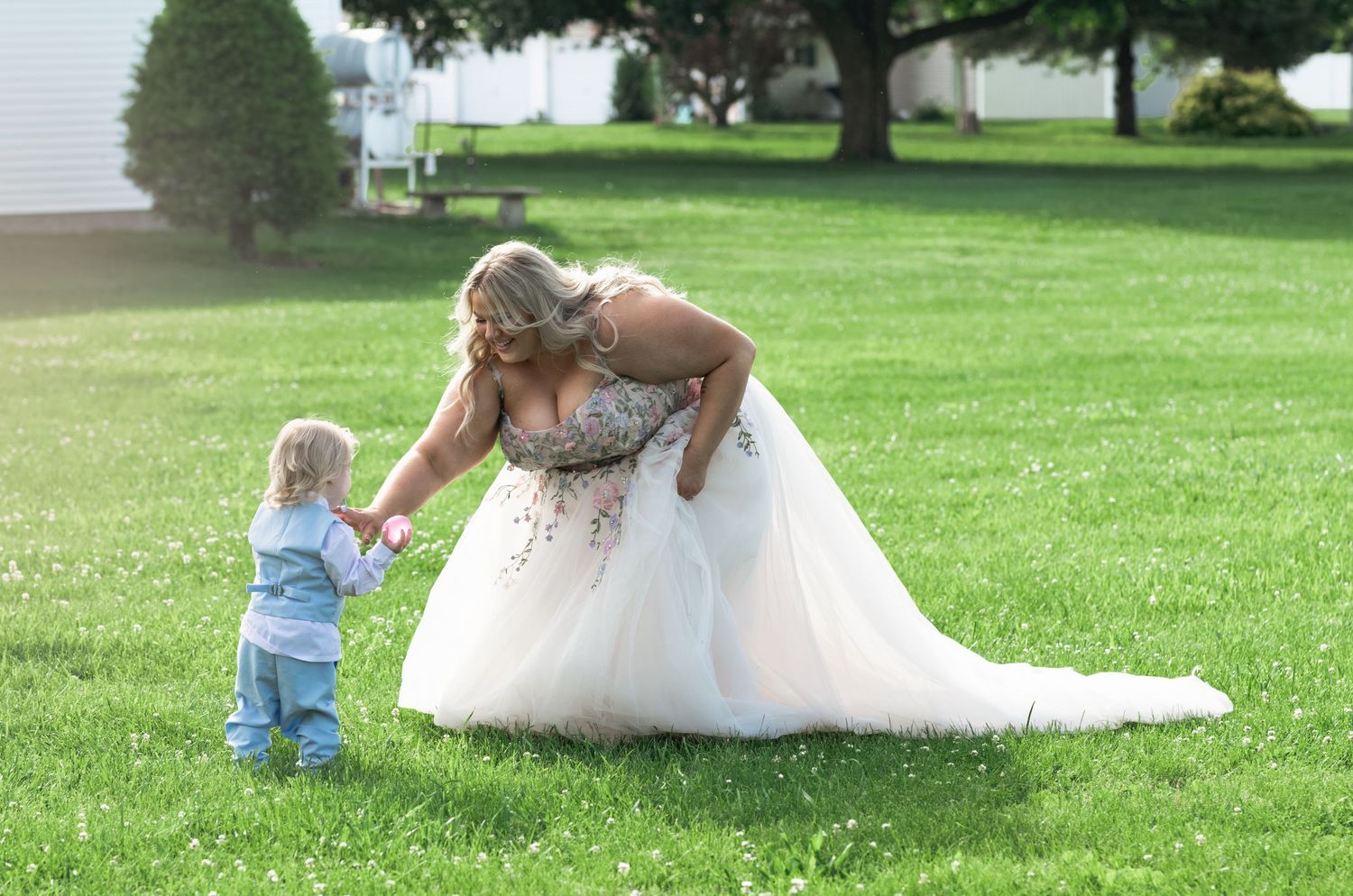 Bride in white ballgown bending down to hold hands with a toddler in blue on a green lawn.