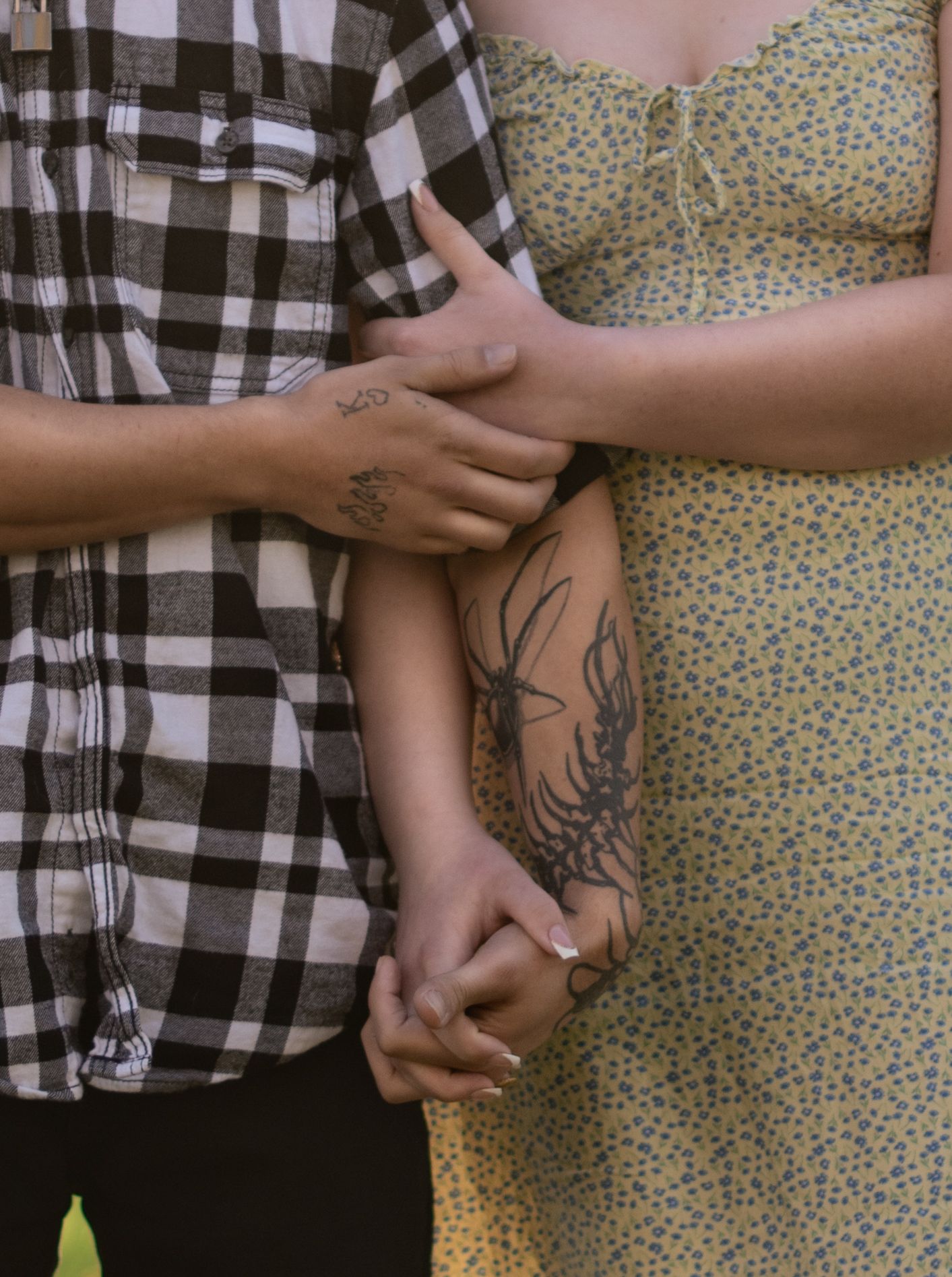 Couple holding hands, woman in floral yellow dress and man in plaid shirt, both with tattoos on their arms.