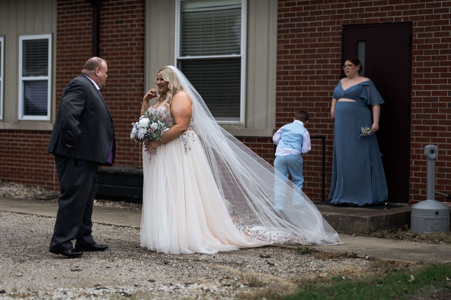 Bride in white gown with veil holds bouquet outside brick church, with bridesmaid in blue dress nearby.