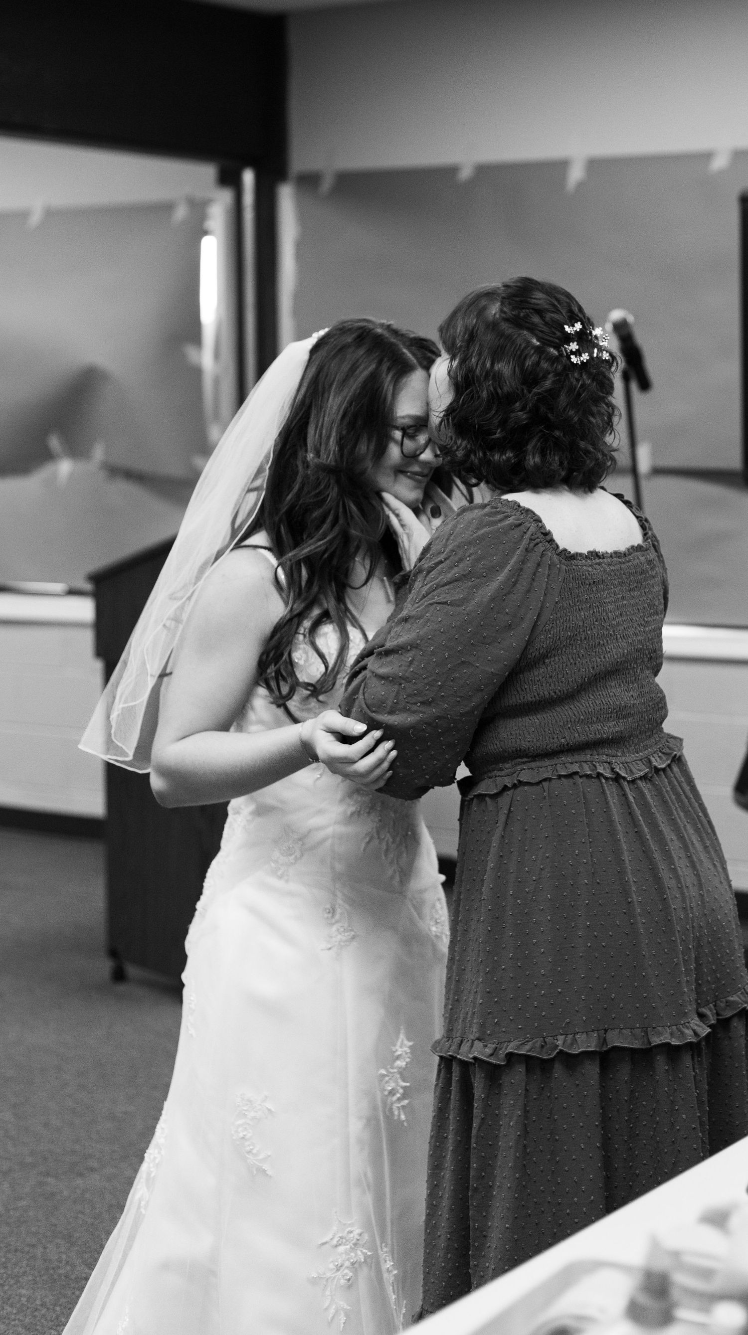 Black and white photo of a bride in a white gown and veil sharing a tender embrace with a woman in a dark dress.