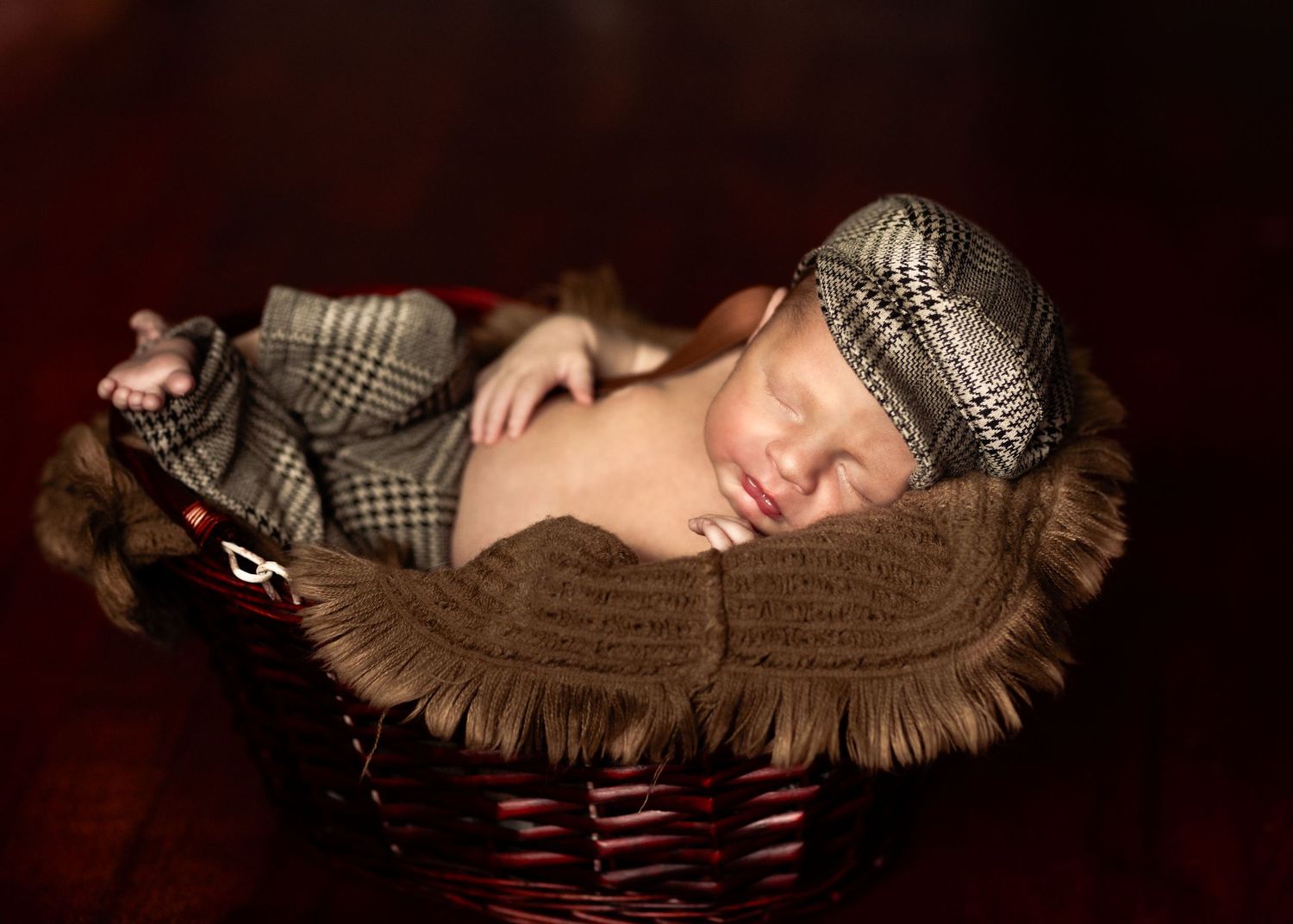 Sleeping newborn baby in plaid newsboy cap and outfit curled in a wicker basket.