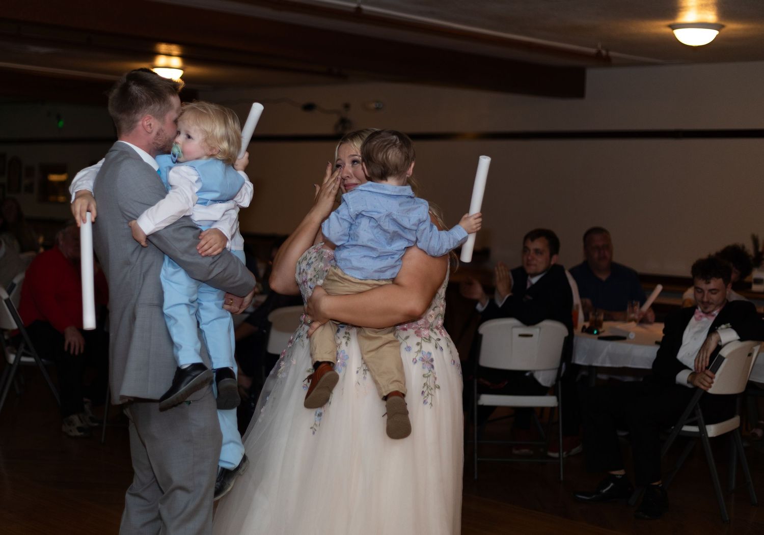 Bride and groom each hold a toddler while dancing at wedding reception with guests seated behind.