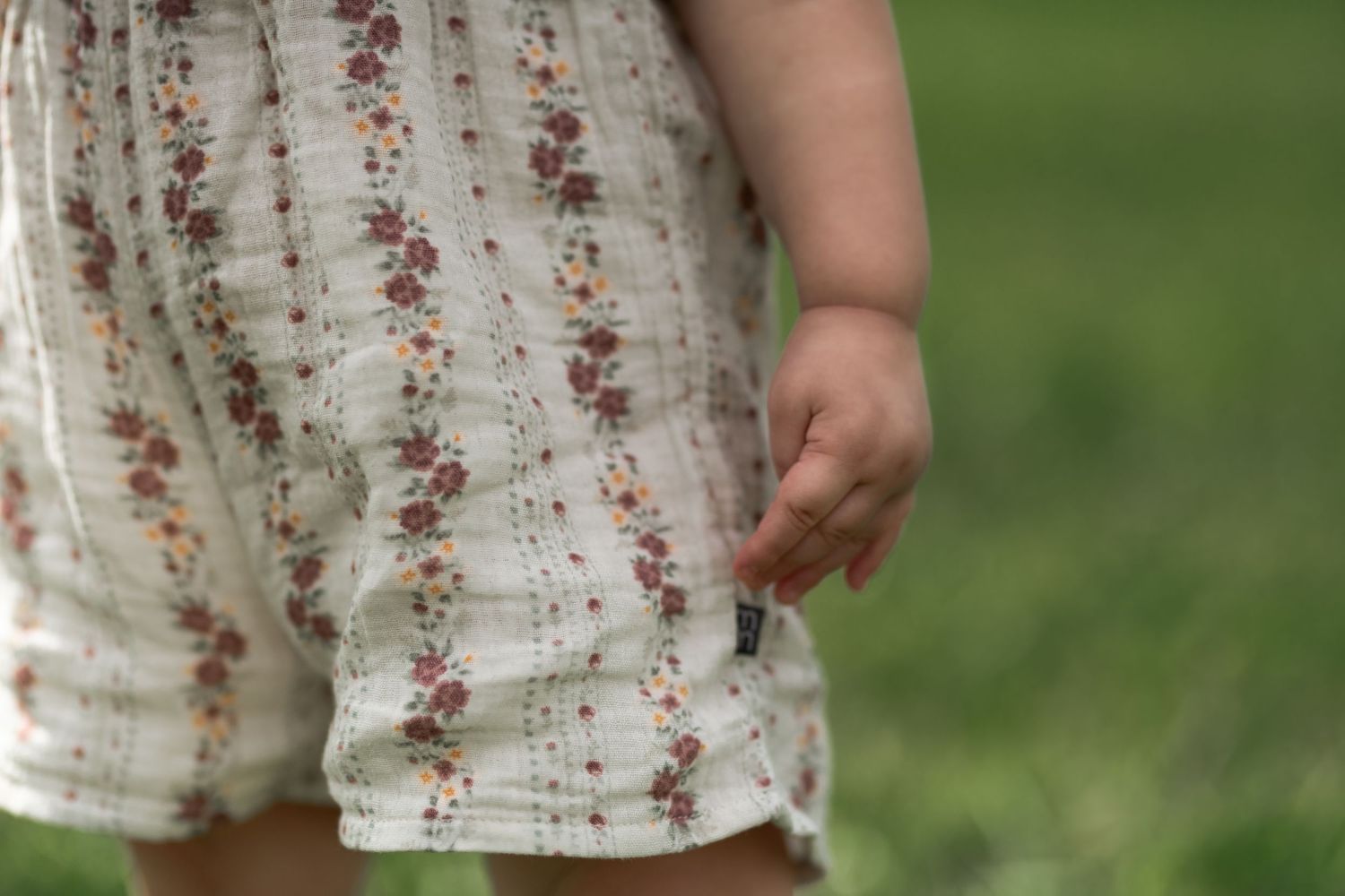 Close-up of a young child wearing a white floral romper, standing on green grass.
