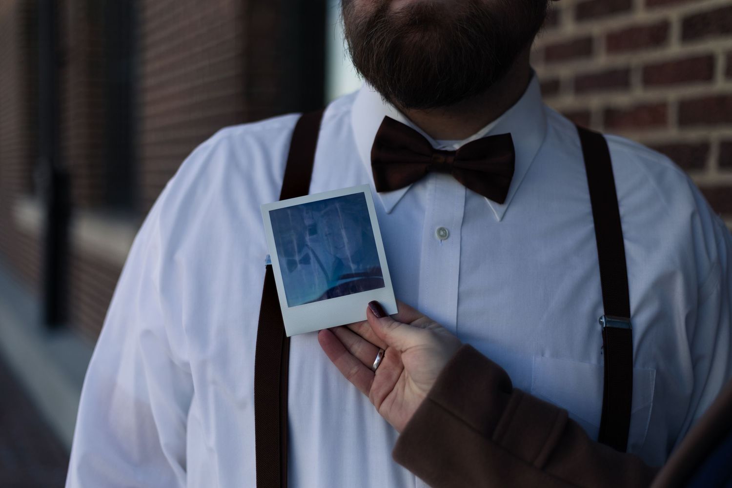 Bearded man in white shirt, bow tie, and suspenders holding a Polaroid photo outdoors near a brick wall.