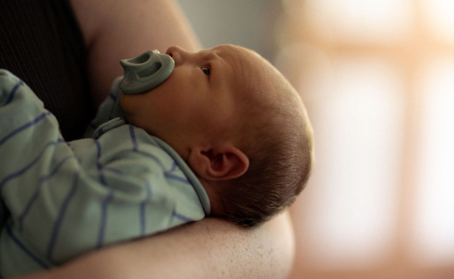 Peaceful newborn baby with pacifier resting in parent's arms in soft natural light.