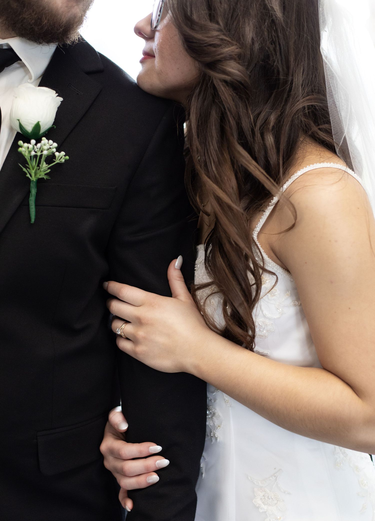 A bride in a white gown and veil embraces a groom in a black suit with a white rose boutonniere on their wedding day.