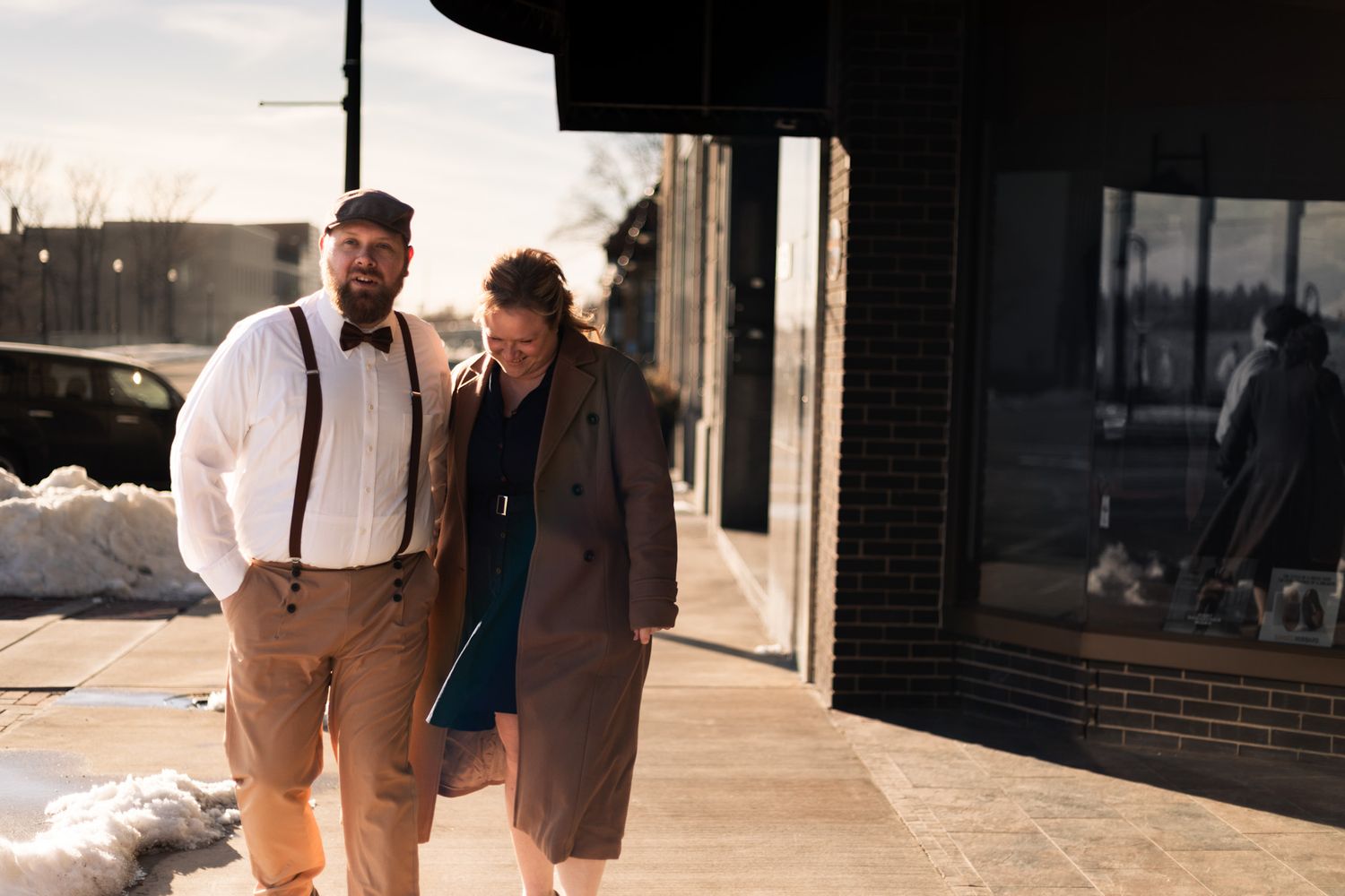 Two people walking together on a sidewalk outside a storefront in winter, dressed in vintage-style clothing.