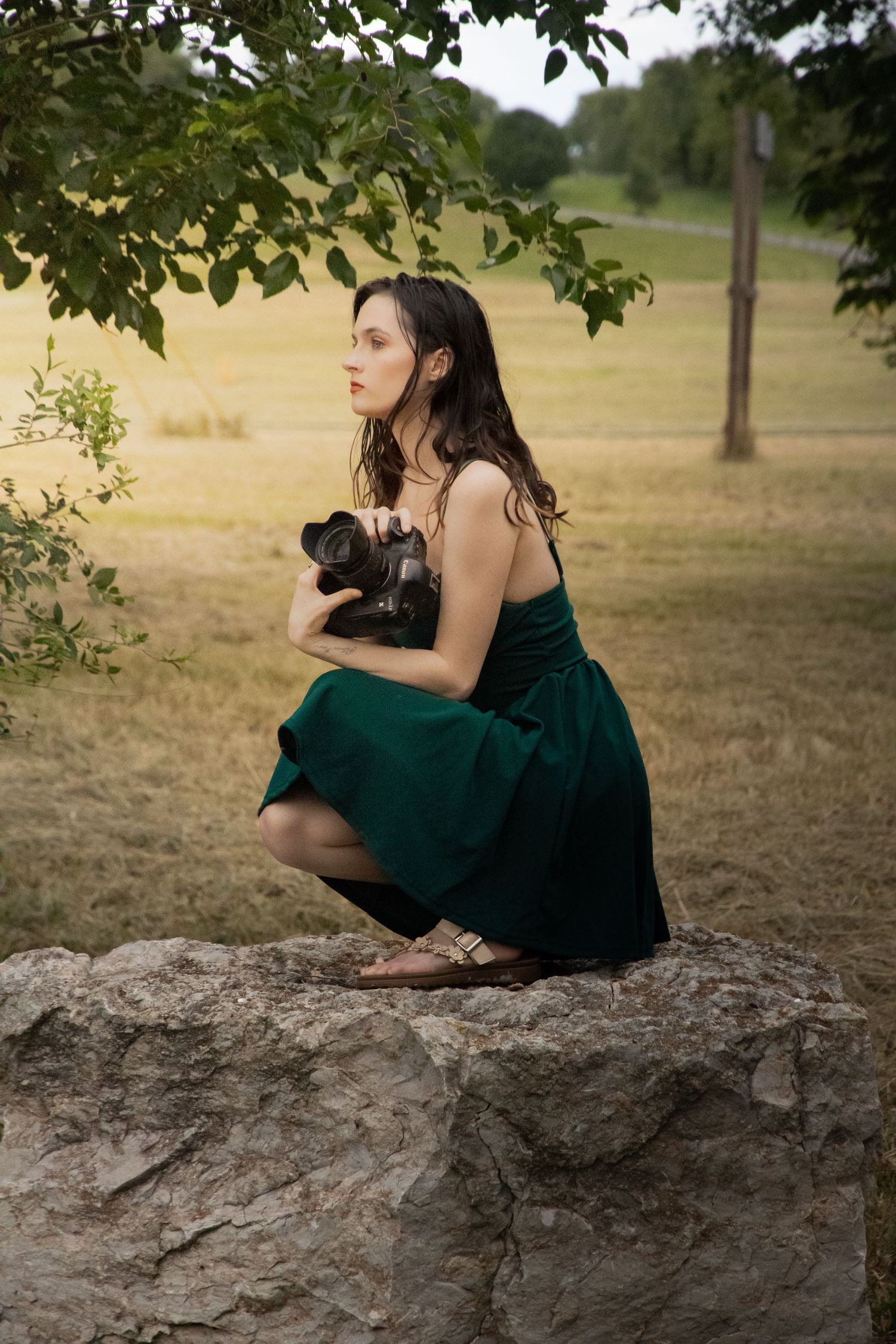 Person in emerald dress sits on large rock with camera in outdoor nature setting.