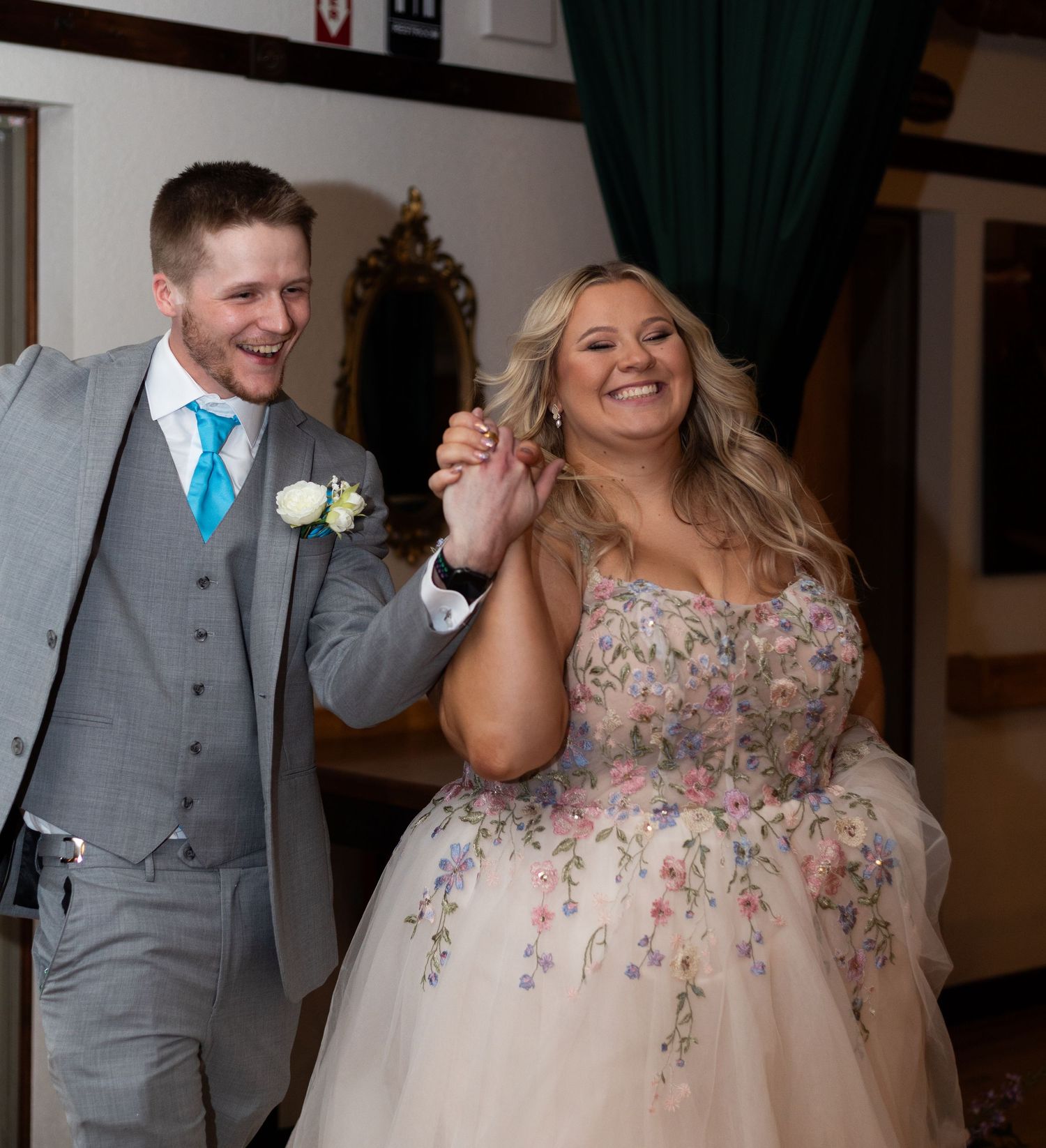 Smiling bride in floral gown holds hands with groom in gray suit and teal tie at wedding reception.