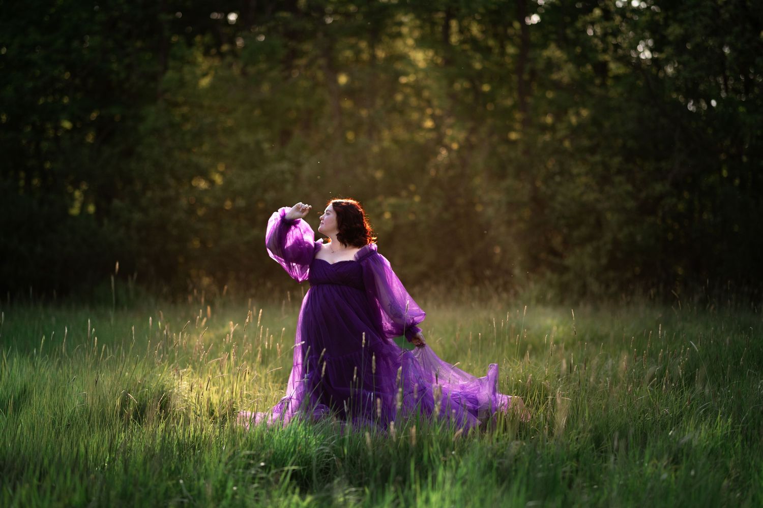 Woman in flowing purple gown poses gracefully in sunlit green meadow surrounded by lush trees.