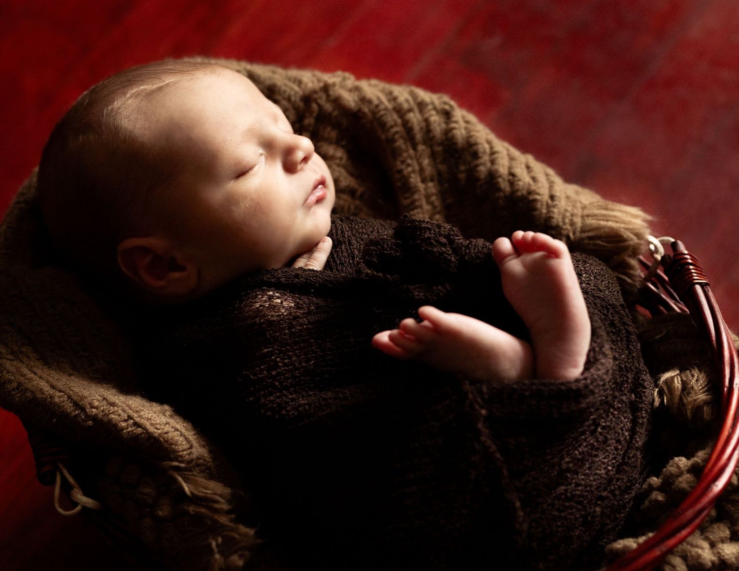 Peaceful newborn baby sleeping in a rustic red basket, wrapped in a soft brown knit blanket on a dark background.