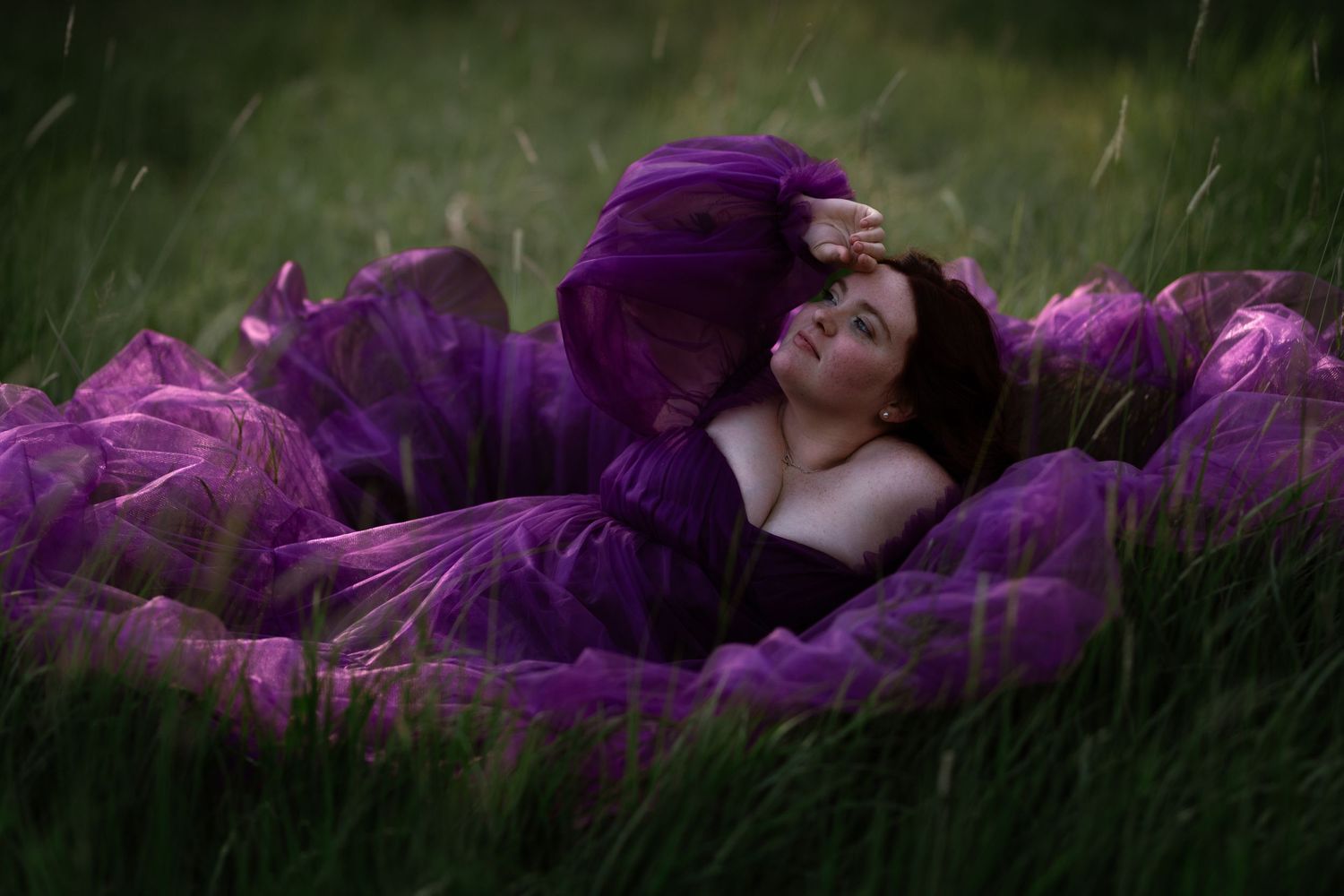 Woman in purple gown lying in green grass field, dreamy artistic portrait photography.