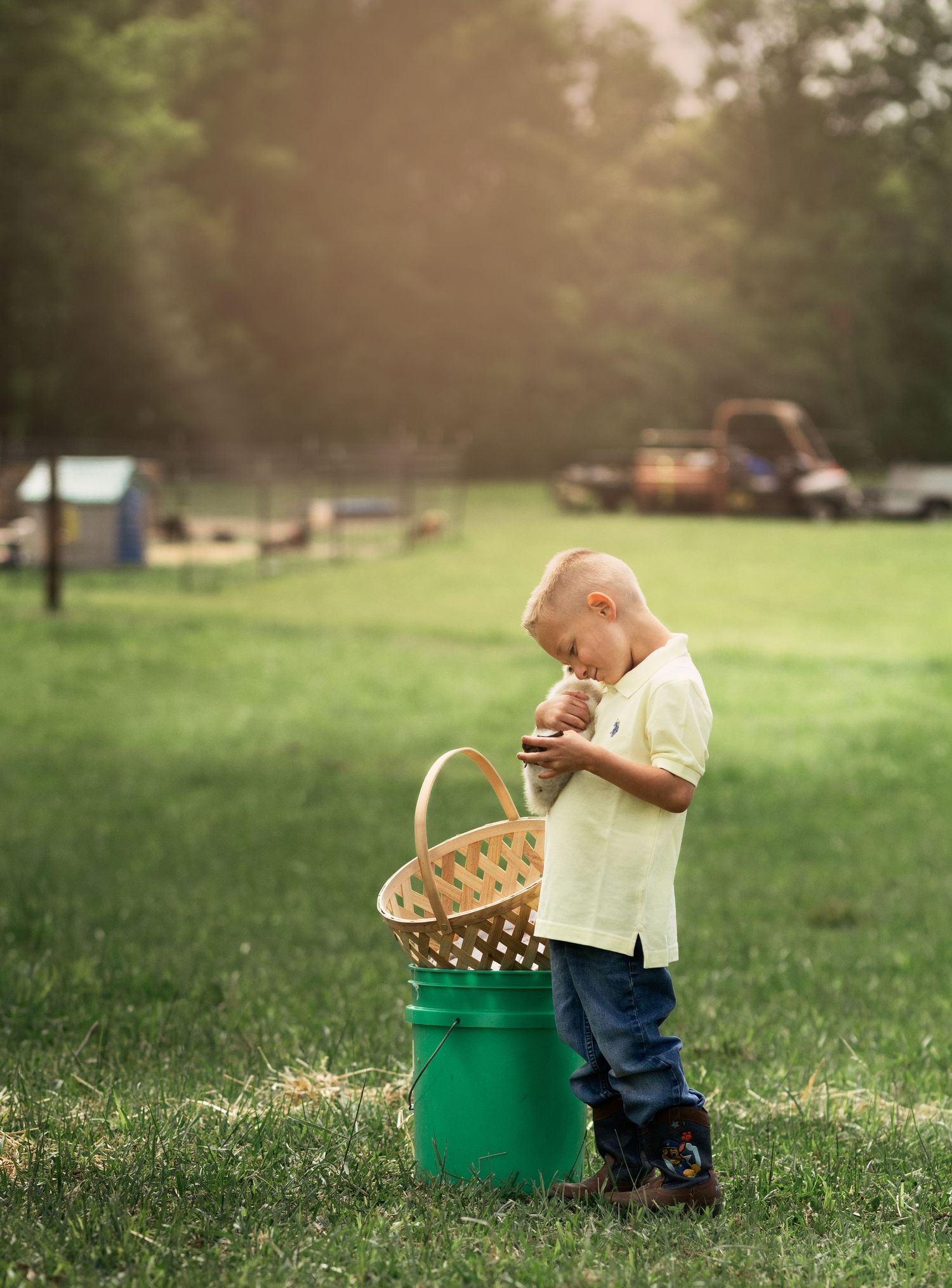 Young boy in yellow shirt holds a chick near a wicker basket on a green bucket outdoors.