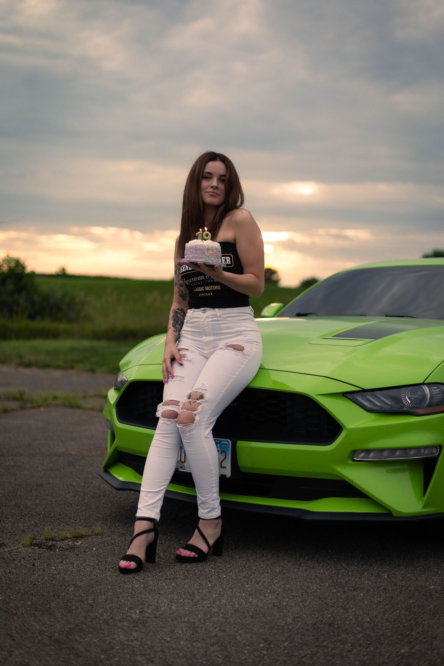 Person in black top and white jeans leans against lime green Mustang at dusk.
