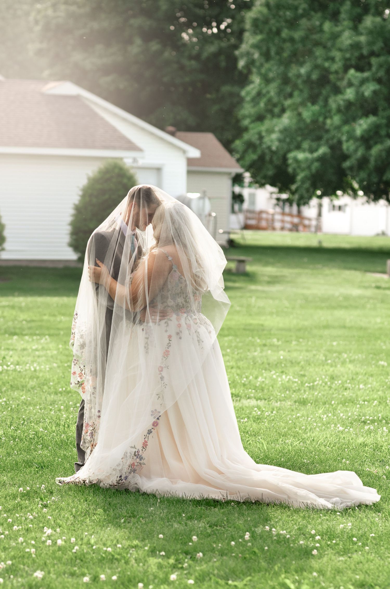 Bride and groom share a romantic kiss under a flowing veil on a lush green lawn at their wedding.