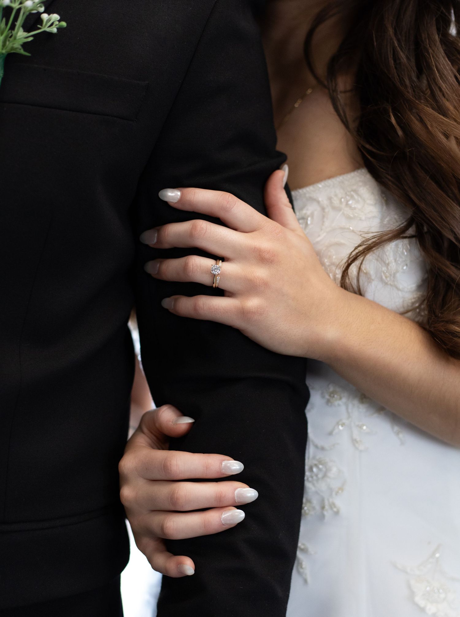 Close-up of a bride's hands with white manicured nails and engagement ring holding onto a black suit jacket.