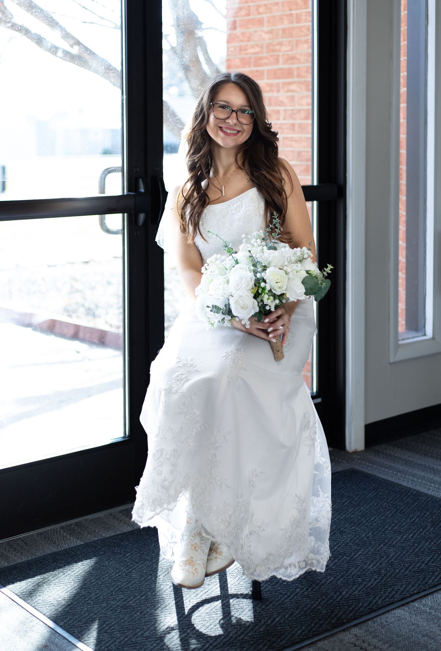 A bride in a white lace dress and glasses holds a small bouquet while standing in a doorway on a snowy day.