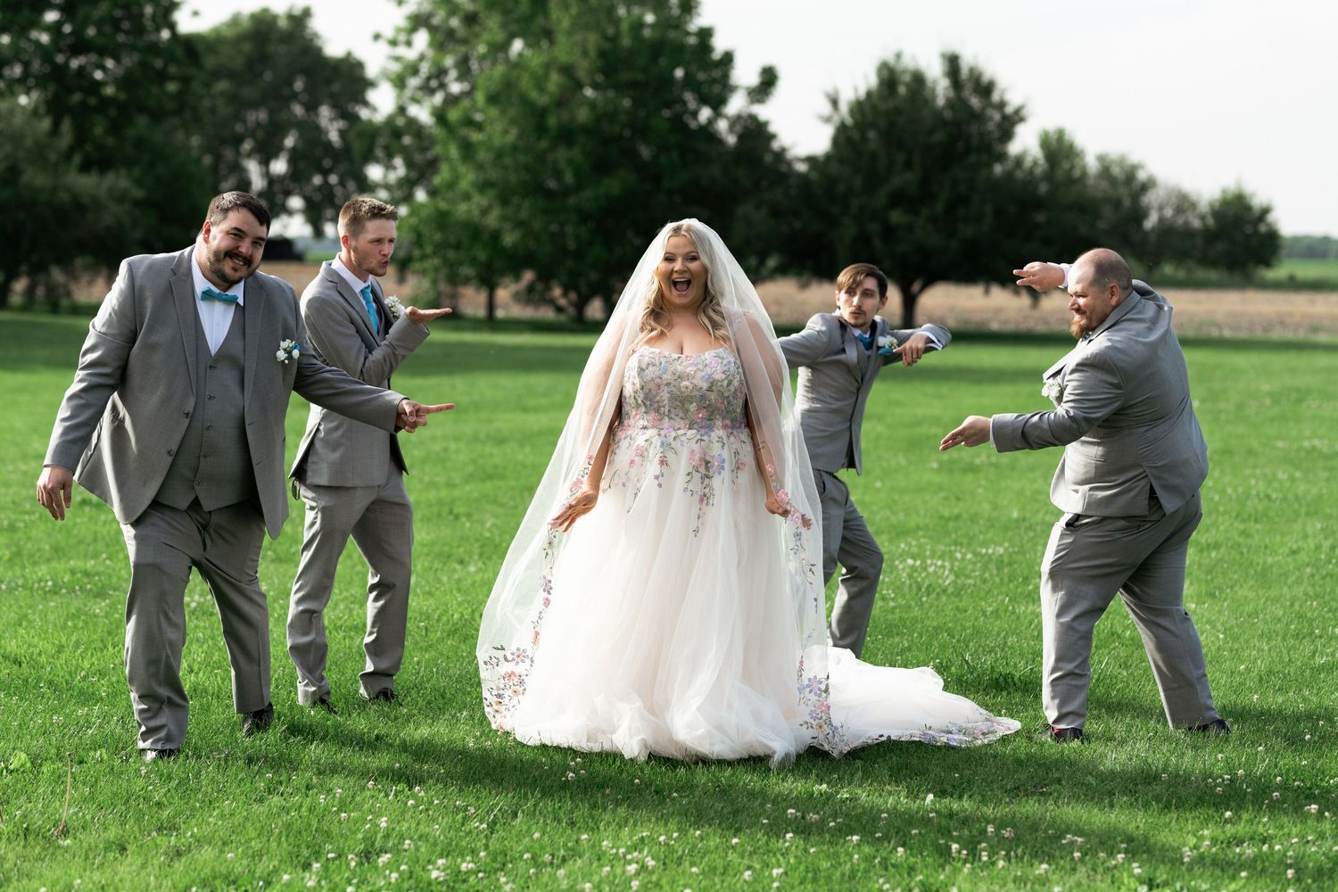 Bride in white ball gown with veil poses on green lawn while groomsmen in gray suits playfully point at her.