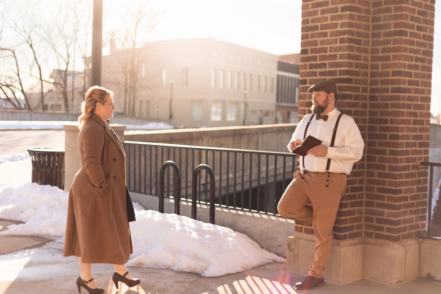 Two people in winter coats and vintage attire posing against a brick building in bright sunlight.