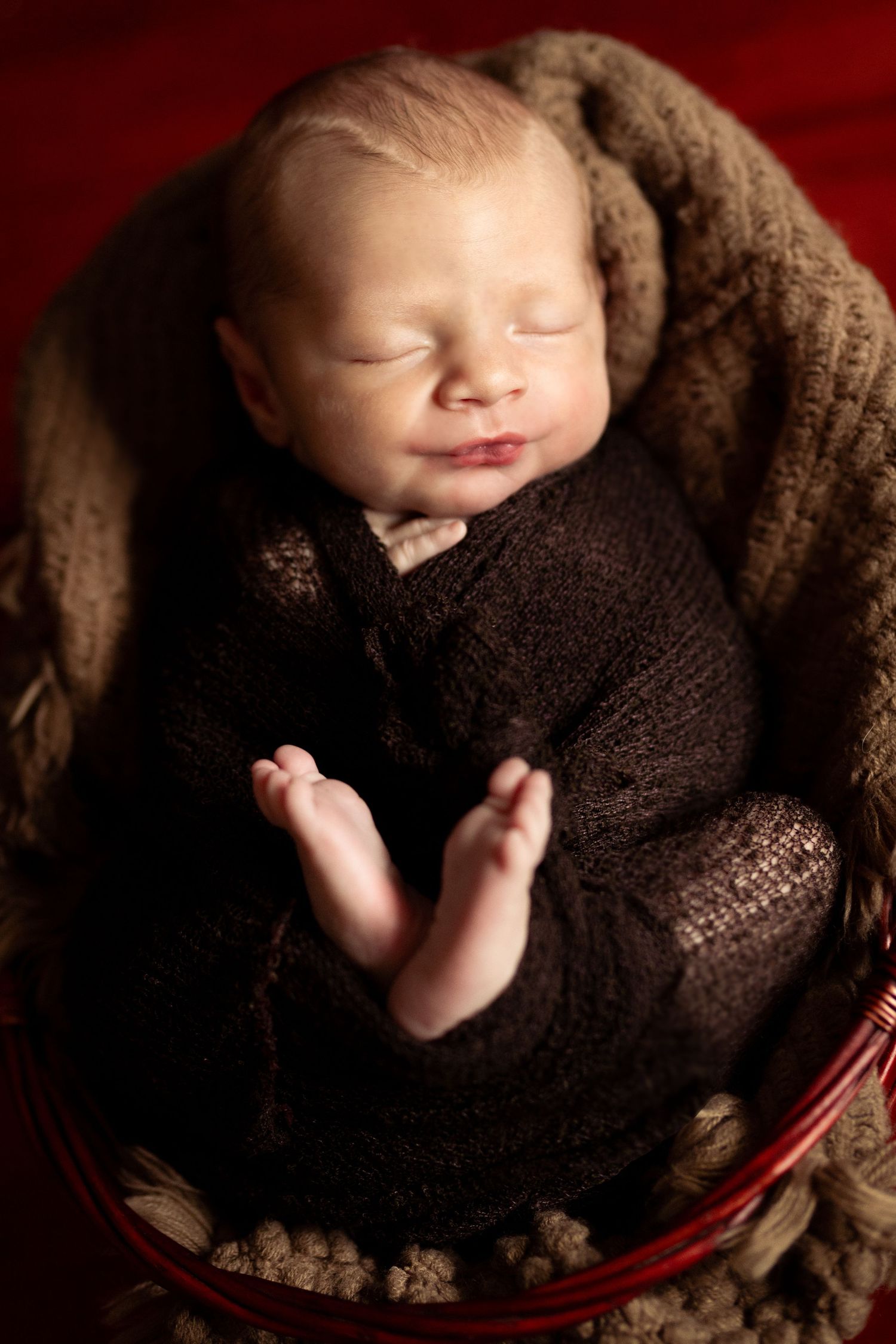 Sleeping newborn baby wrapped in dark brown knit swaddle nestled in a rustic red basket with soft blanket.