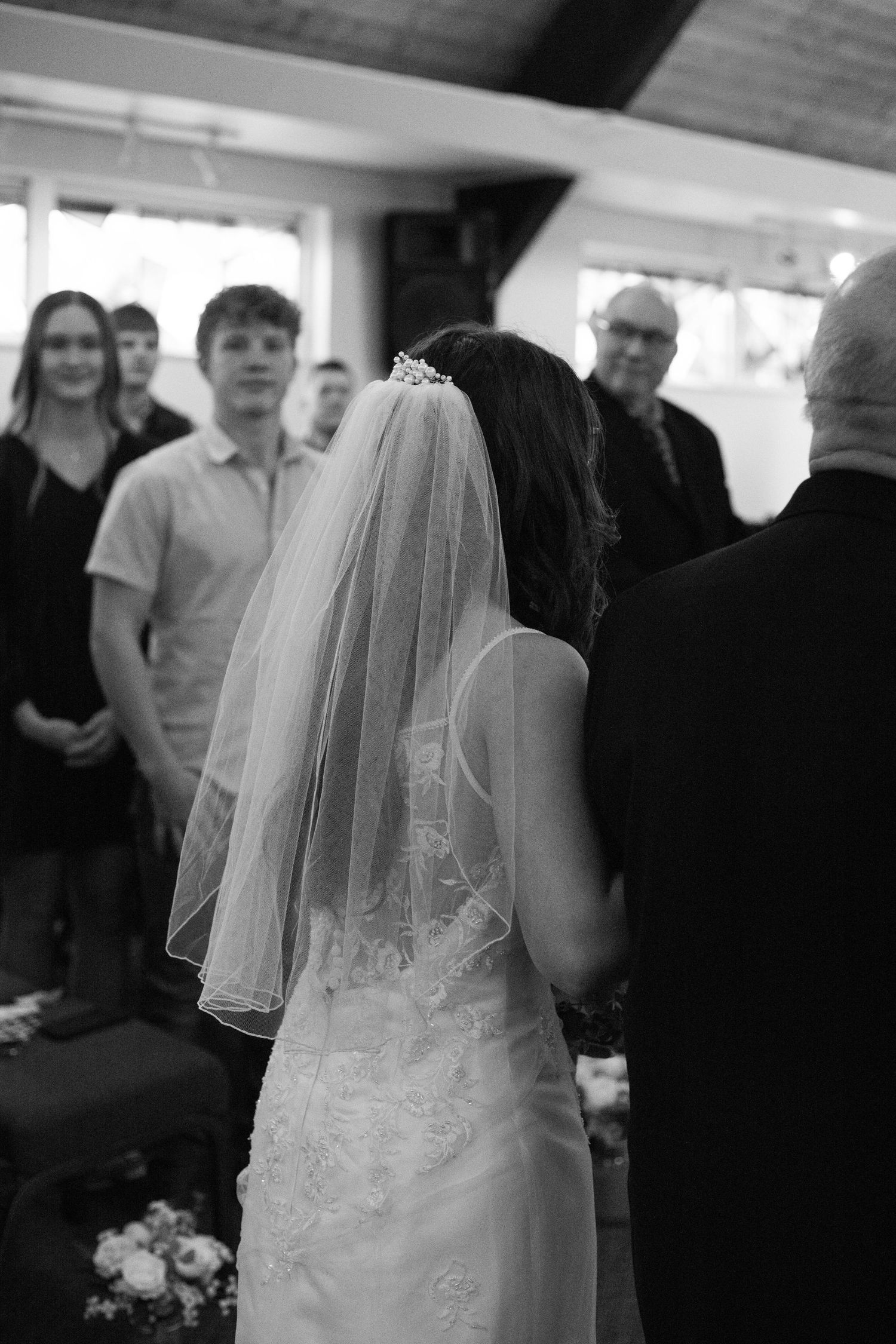 Black and white photo of a bride in a lace gown and veil at her wedding ceremony with guests watching.