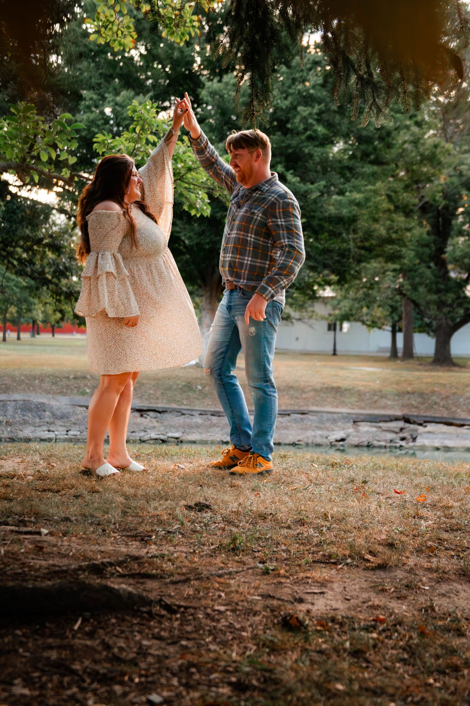 A couple stands together in a park during golden hour, with the person in a flowing dress and plaid shirt.