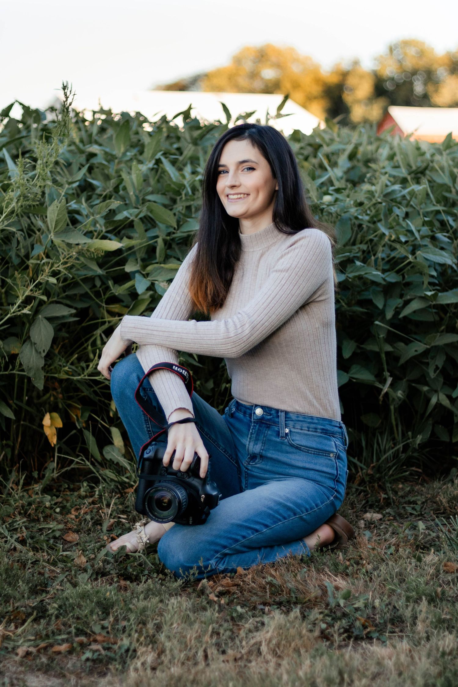 Woman in beige sweater and jeans relaxes in a garden setting during a casual photoshoot.