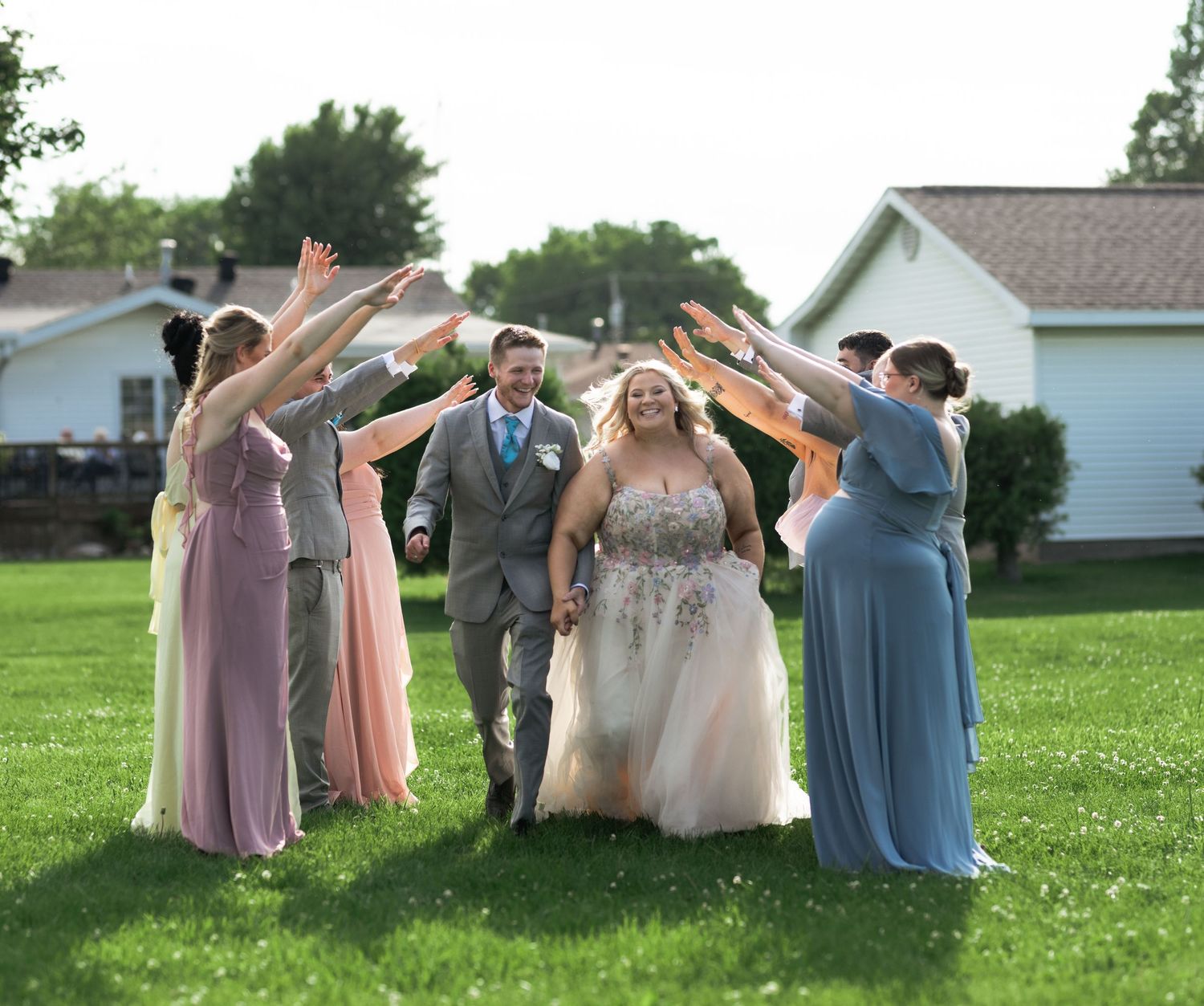 Bride and groom walk under arch of raised hands formed by bridesmaids in colorful gowns on green lawn.