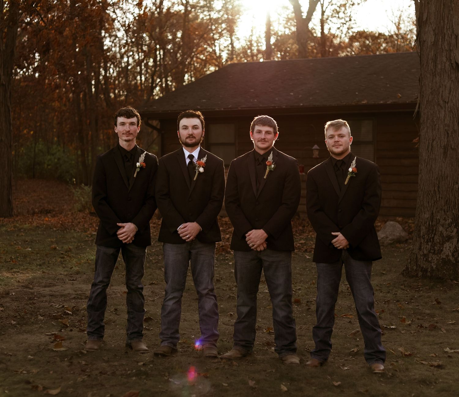 A group of people in formal black attire stand together at sunset in a wooded outdoor setting.