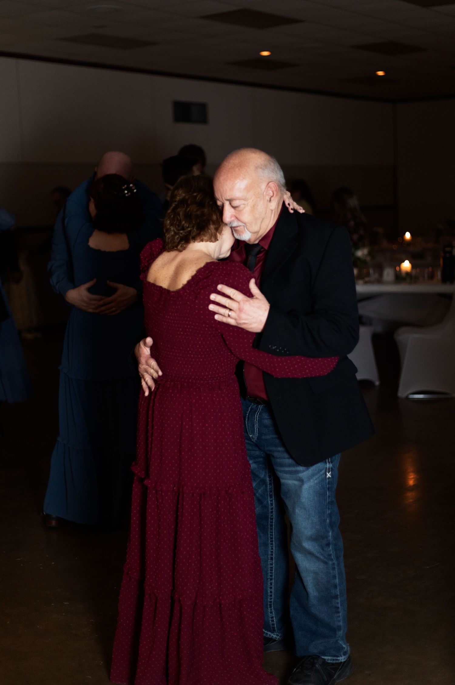 A couple dances together in a dimly lit room, the woman wearing a burgundy dress and her partner in dark clothing.