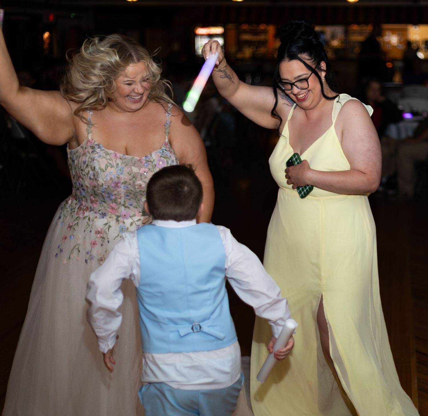 Two women in formal gowns dancing with a young boy at a wedding reception, holding glow sticks.