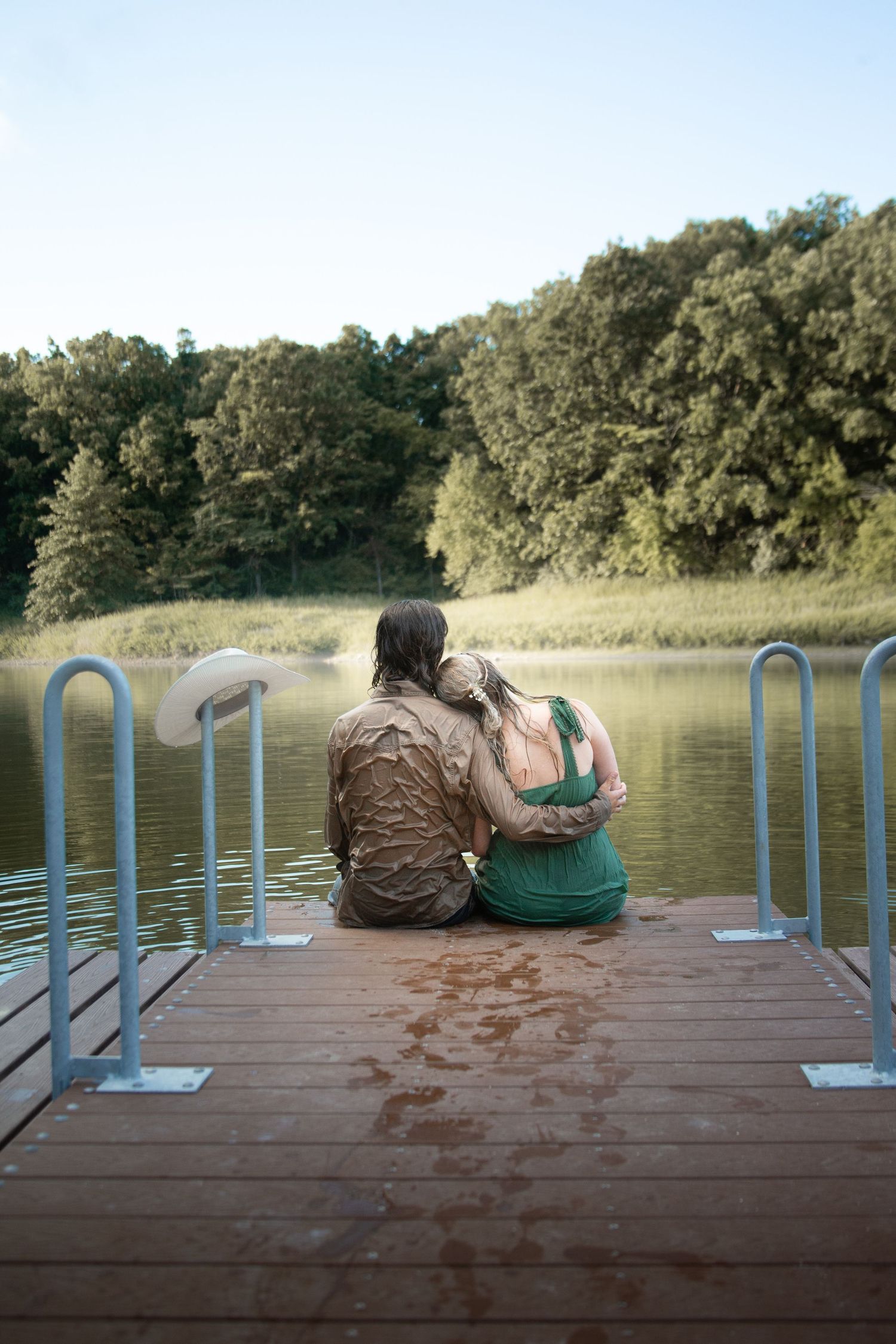 Two people sit together on a wooden dock overlooking a peaceful lake surrounded by trees.