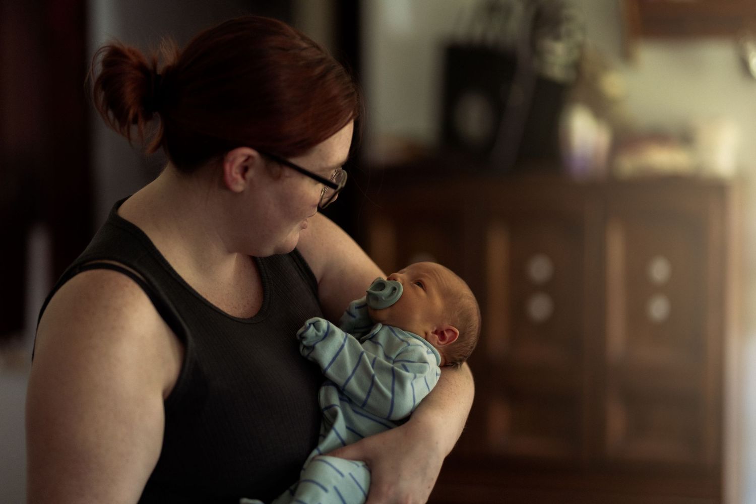 A mother with red hair and glasses tenderly holds her newborn baby in a softly lit room.