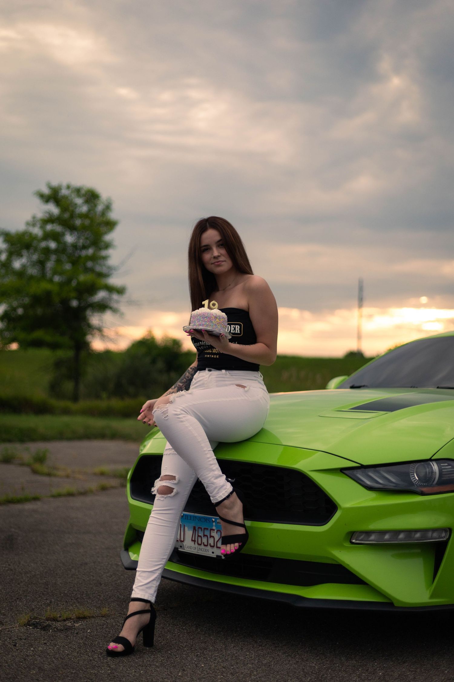 Person in white outfit poses on bright green Mustang sports car at sunset.
