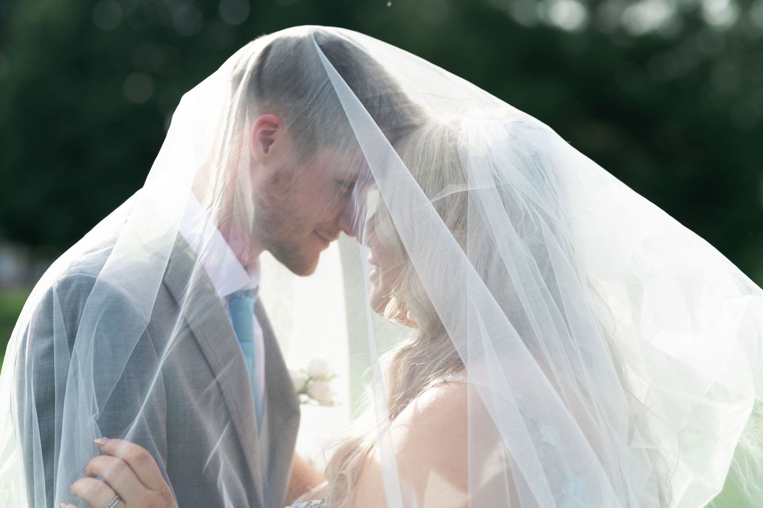 Bride and groom share an intimate moment under a flowing white wedding veil outdoors on their wedding day.