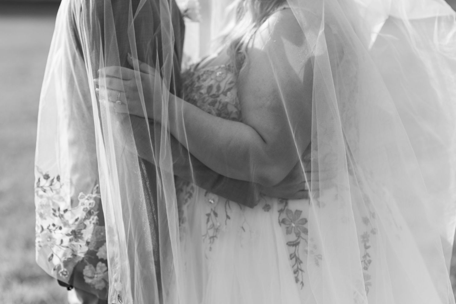 Black and white photo of two brides embracing under a flowing veil on their wedding day.