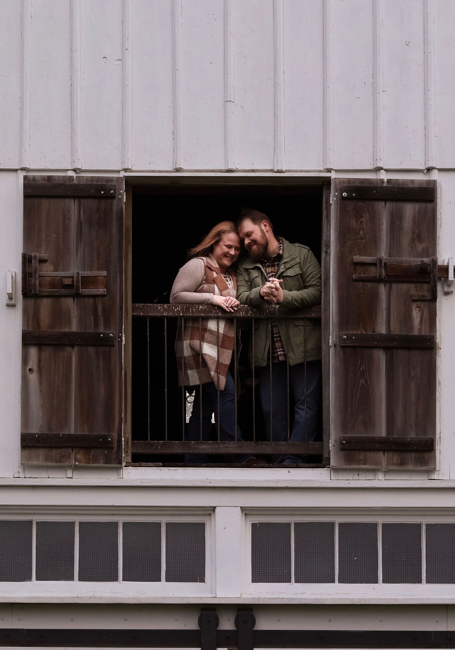 A couple poses playfully in the window frame of a white barn building.